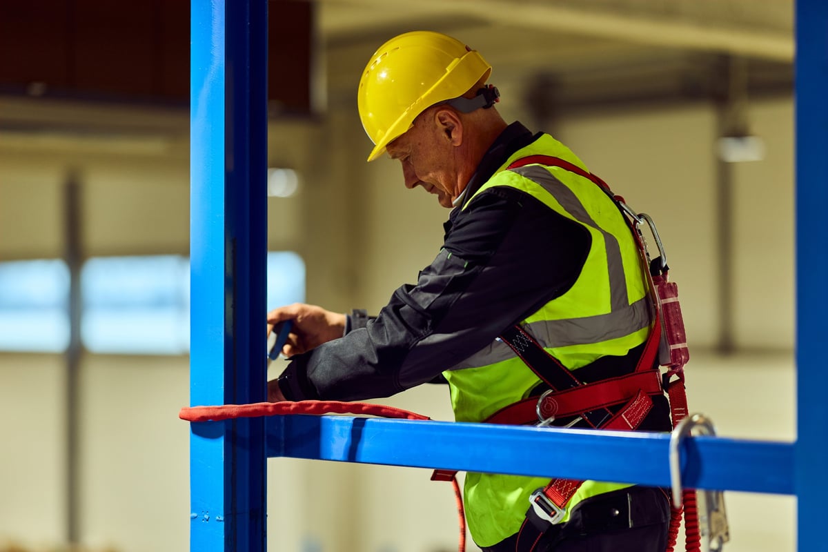 Male construction worker in a warehouse wearing a yellow hard hat, hi-vis vest and safety harness while inspecting and securing a blue scaffold