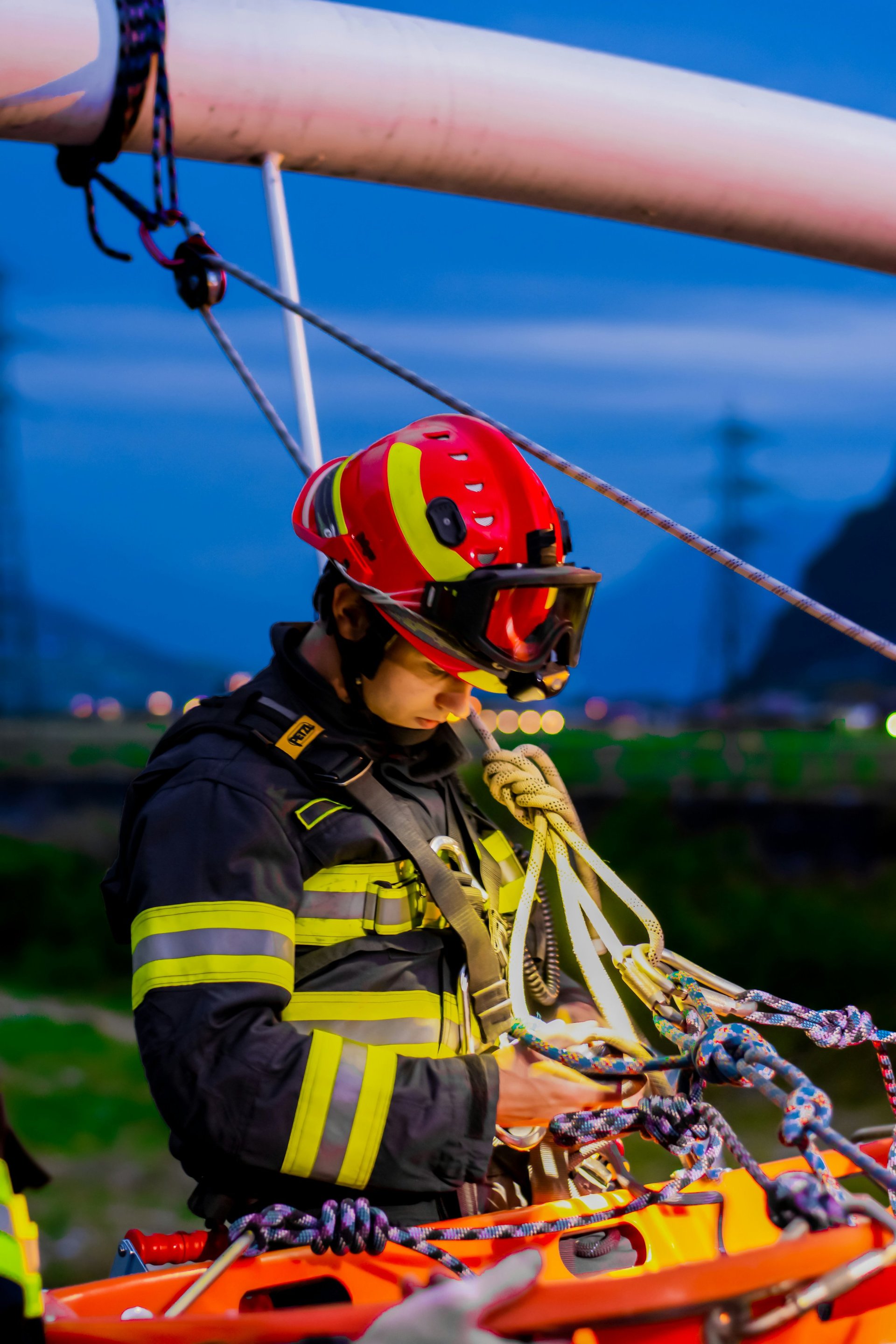 Feuerwehrmann in rotem Helm und dunkler Uniform mit reflektierenden Streifen hält Seil bei Rettungsübung gegen blauen Himmel